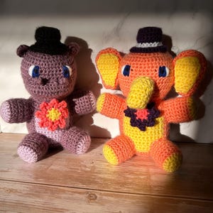 Puede incluir: Dos animales de peluche de ganchillo hechos a mano: un oso morado y un elefante naranja. El oso tiene un sombrero de copa negro y una flor en el pecho. El elefante tiene un sombrero de copa morado y grandes orejas amarillas. Ambos tienen ojos azules.