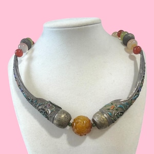 May include: An antique necklace with a silver-tone metal collar and colorful gemstone beads. The necklace features ornate, curved metal pieces with floral and enamel details, connected by a central amber-colored bead and smaller, multi-colored beads.