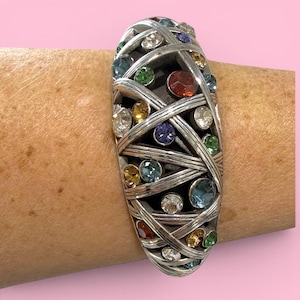 Puede incluir: Brazalete plateado con un dise&ntilde;o tejido, adornado con una variedad de gemas de colores. Las gemas incluyen piedras transparentes, rojas, azules, verdes y amarillas. El brazalete tiene forma ovalada y se usa en la mu&ntilde;eca.