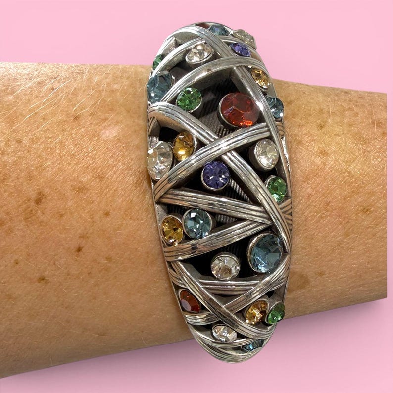 Puede incluir: Brazalete plateado con un dise&ntilde;o tejido, adornado con una variedad de gemas de colores. Las gemas incluyen piedras transparentes, rojas, azules, verdes y amarillas. El brazalete tiene forma ovalada y se usa en la mu&ntilde;eca.