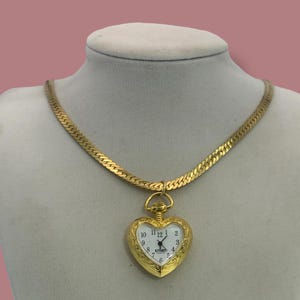 May include: A gold-toned necklace with a heart-shaped pendant watch. The watch face is white with black numerals and hands. The necklace has a flat, herringbone chain design.
