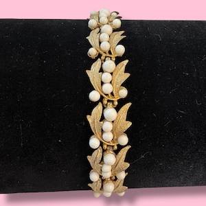 May include: Gold-tone bracelet featuring white faux pearls and leaf-shaped accents. The bracelet has a clasp closure and is displayed on a black surface. The design is vintage-inspired, with a classic and elegant aesthetic.