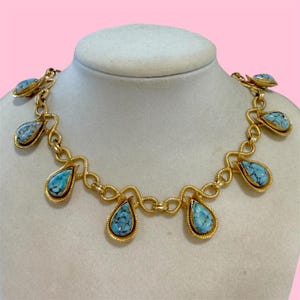 May include: A gold-toned necklace with teardrop-shaped turquoise-colored stones. The necklace is displayed on a mannequin. The stones have a marbled appearance and are set in gold-colored bezels. The chain has a unique, looped design.