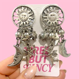 May include: Silver-toned celestial earrings featuring sun and moon charms, stars, and cherubs. The earrings have a dangling design with intricate details. The text on the card reads "FREE BUT FANCY".