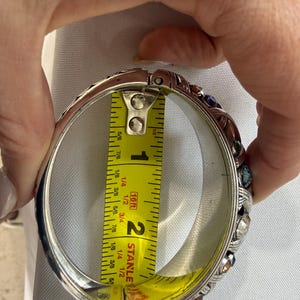 Puede incluir: Brazalete de plata con bisagras y gemas de colores. El brazalete se mide con una cinta m&eacute;trica amarilla, que muestra un di&aacute;metro aproximado de 5 cm. El brazalete tiene un dise&ntilde;o ornamentado con piedras de varios colores.