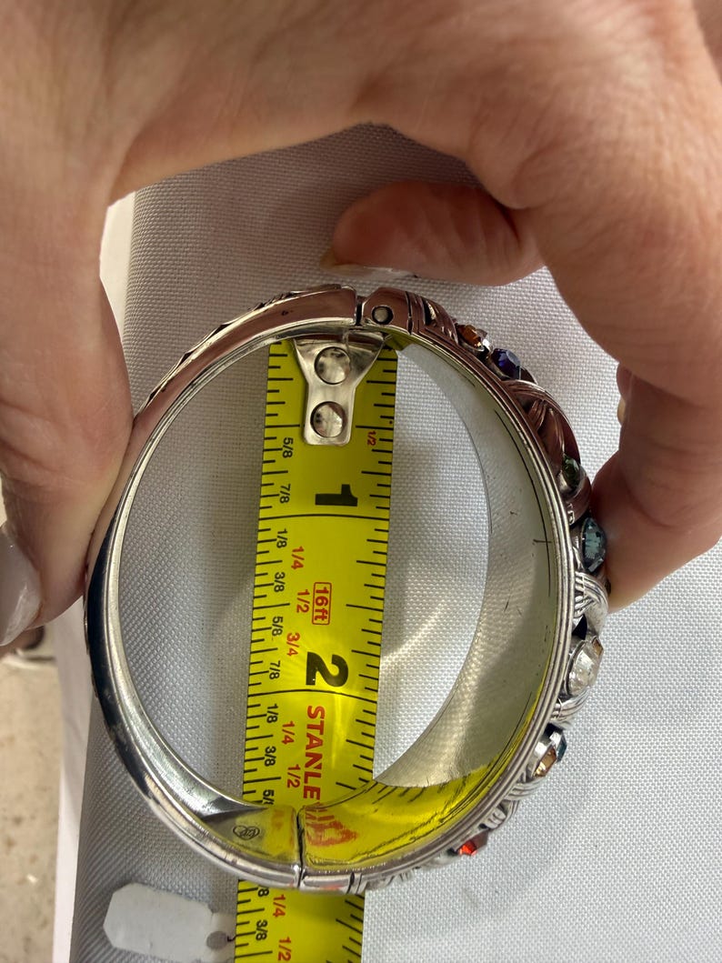 Puede incluir: Brazalete de plata con bisagras y gemas de colores. El brazalete se mide con una cinta m&eacute;trica amarilla, que muestra un di&aacute;metro aproximado de 5 cm. El brazalete tiene un dise&ntilde;o ornamentado con piedras de varios colores.