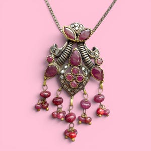 May include: A silver-toned pendant necklace with a floral design, featuring ruby-colored gemstones and small clear stones. The pendant has a detailed, ornate design with dangling ruby beads. The background is a solid pink.