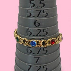 May include: A gold-toned bracelet with a chain link design, adorned with colorful gemstones. The bracelet is displayed on a gray ring sizer, showcasing its size. The gemstones are green, blue, and red.