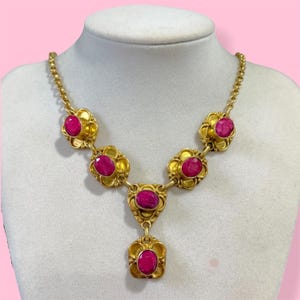 May include: A gold-toned necklace with a series of ruby-colored gemstones set in ornate floral settings. The necklace is displayed on a white mannequin bust against a pink background. The gemstones are oval and round, and the necklace has a delicate chain.