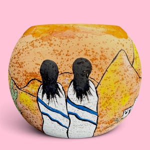 May include: A round ceramic vase featuring a desert landscape painting with two figures in white cloaks with blue stripes, black hair, a green cactus, and yellow mountains against a speckled orange and tan background.