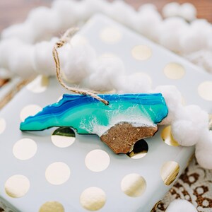 May include: A wooden ornament in the shape of North Carolina, featuring blue and green resin representing the ocean and a sandy beach. The ornament is on a wrapped gift with gold polka dots, with a white pom-pom garland.