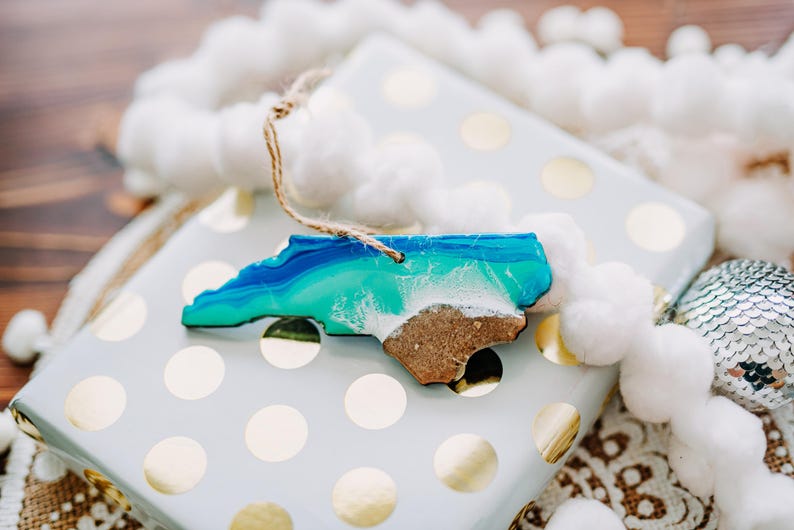 May include: A wooden ornament in the shape of North Carolina, featuring blue and green resin representing the ocean and a sandy beach. The ornament is on a wrapped gift with gold polka dots, with a white pom-pom garland.