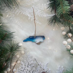 May include: A wooden ornament in the shape of North Carolina, with blue and white resin representing the ocean and sky. The ornament hangs from a twine string, surrounded by Christmas greenery, white berries, and pinecones.