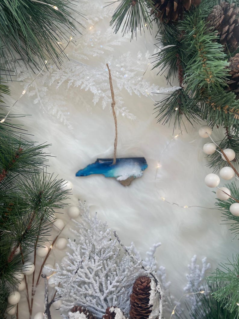 May include: A wooden ornament in the shape of North Carolina, with blue and white resin representing the ocean and sky. The ornament hangs from a twine string, surrounded by Christmas greenery, white berries, and pinecones.
