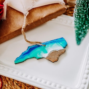May include: A decorative ornament in the shape of North Carolina, featuring a blue and turquoise ocean design with a white foamy wave and a brown sand section. It has a jute string for hanging.