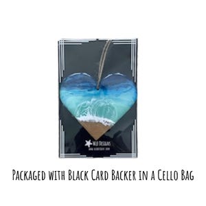 May include: Heart-shaped ornament featuring a beach scene with blue and turquoise waves, a sandy shore, and a jute string for hanging. The ornament is packaged on a black card with white border details and the text "WILD DESIGNS".