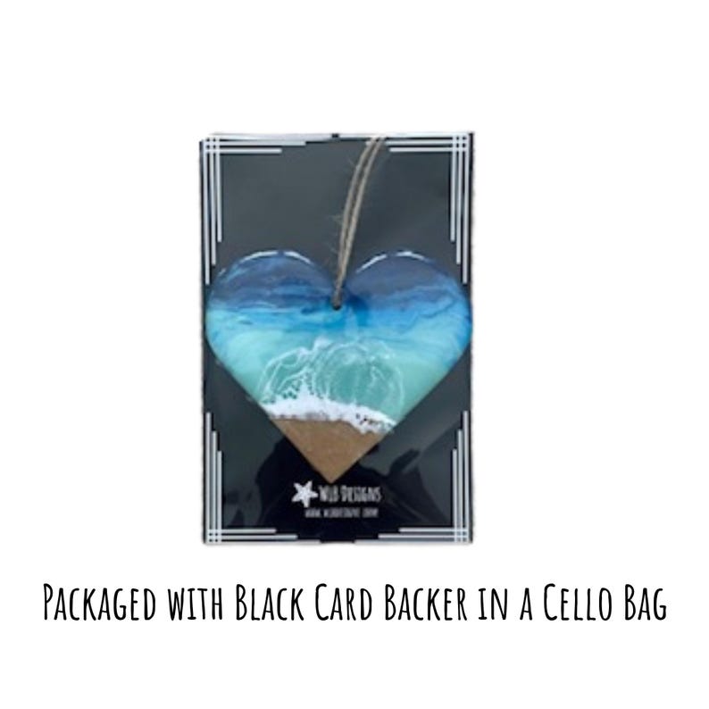 May include: Heart-shaped ornament featuring a beach scene with blue and turquoise waves, a sandy shore, and a jute string for hanging. The ornament is packaged on a black card with white border details and the text "WILD DESIGNS".