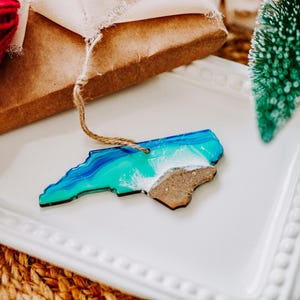 May include: A North Carolina state-shaped ornament with a blue and green ocean design and a brown wooden landmass. The ornament is on a white tray with a small green Christmas tree and a ball of red yarn.