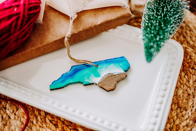 May include: A North Carolina state-shaped ornament with a blue and green ocean design and a brown wooden landmass. The ornament is on a white tray with a small green Christmas tree and a ball of red yarn.