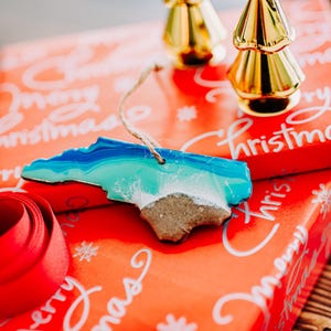 May include: A North Carolina-shaped ornament with blue, turquoise, and white resin, resembling ocean waves and a sandy beach. It hangs by a twine string. Resting on a red gift box with "Merry Christmas" text and a red ribbon.