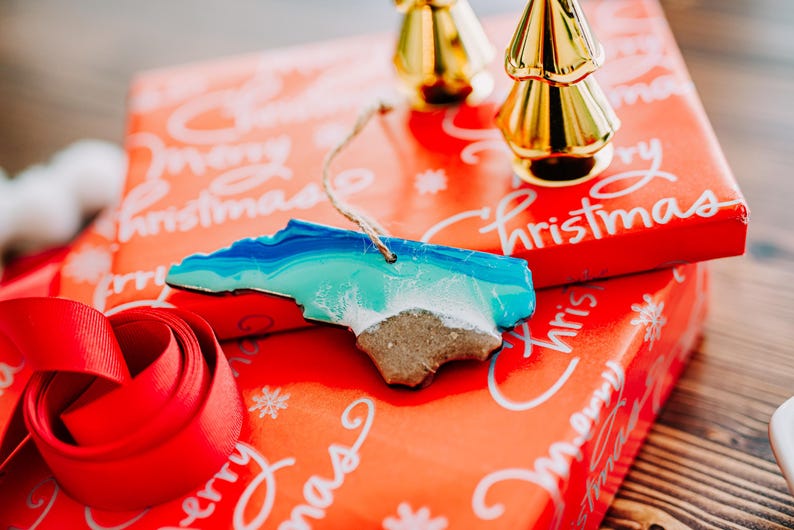 May include: A North Carolina-shaped ornament with blue, turquoise, and white resin, resembling ocean waves and a sandy beach. It hangs by a twine string. Resting on a red gift box with "Merry Christmas" text and a red ribbon.