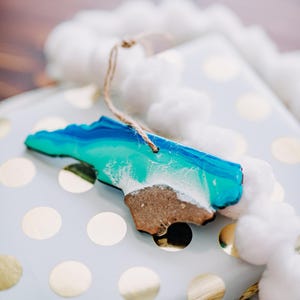 May include: A wooden ornament in the shape of a state, featuring a beach and ocean design in shades of blue, teal, and white. The ornament has a brown sand-like base and a string for hanging. It rests on a white gift box with gold polka dots.