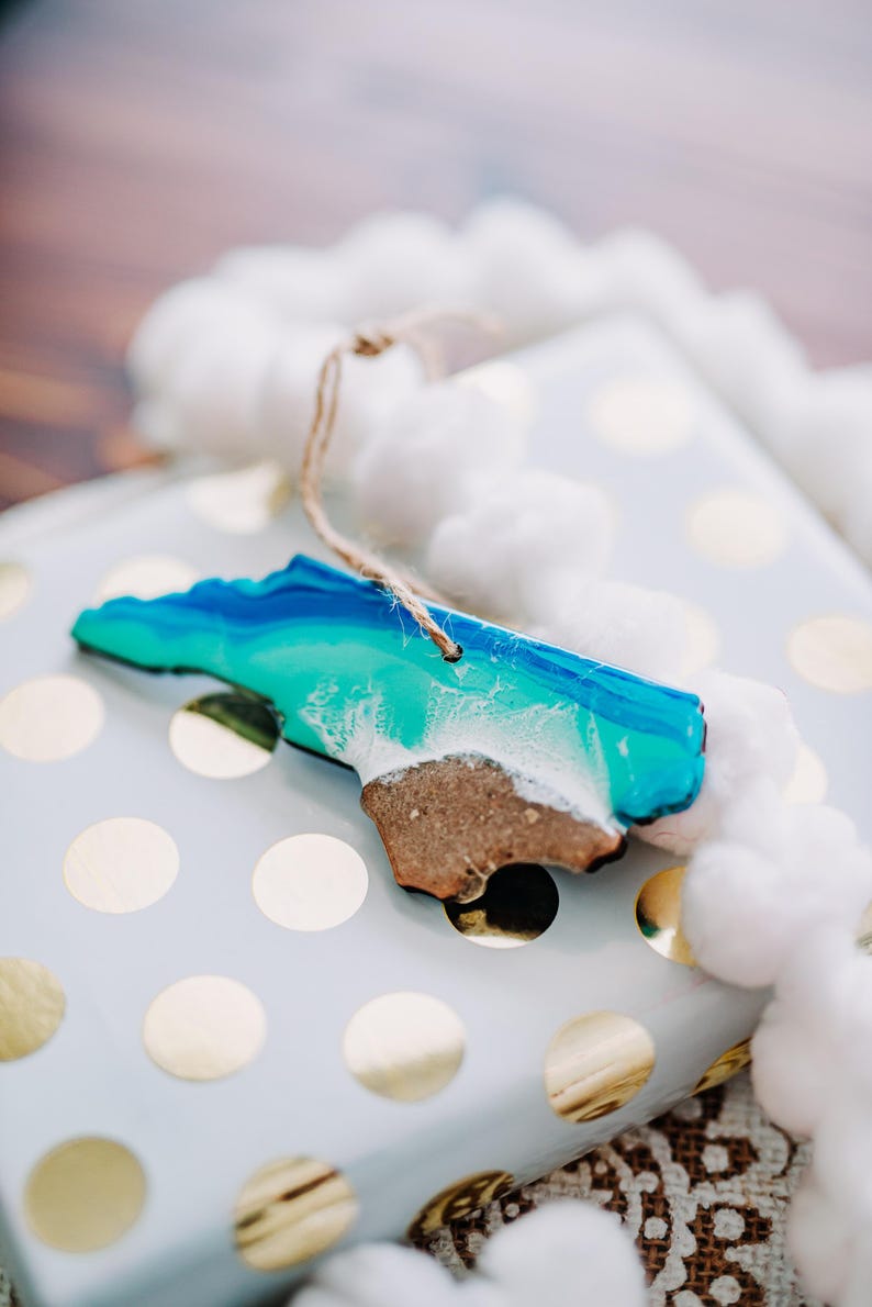 May include: A wooden ornament in the shape of a state, featuring a beach and ocean design in shades of blue, teal, and white. The ornament has a brown sand-like base and a string for hanging. It rests on a white gift box with gold polka dots.