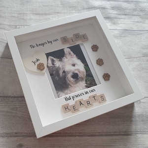 May include: White framed photo memorial with a picture of a white dog. The frame has a heart with the name "Josh" and paw prints. The text "No longer by our side" and "But forever in our hearts" is spelled out with letter tiles.