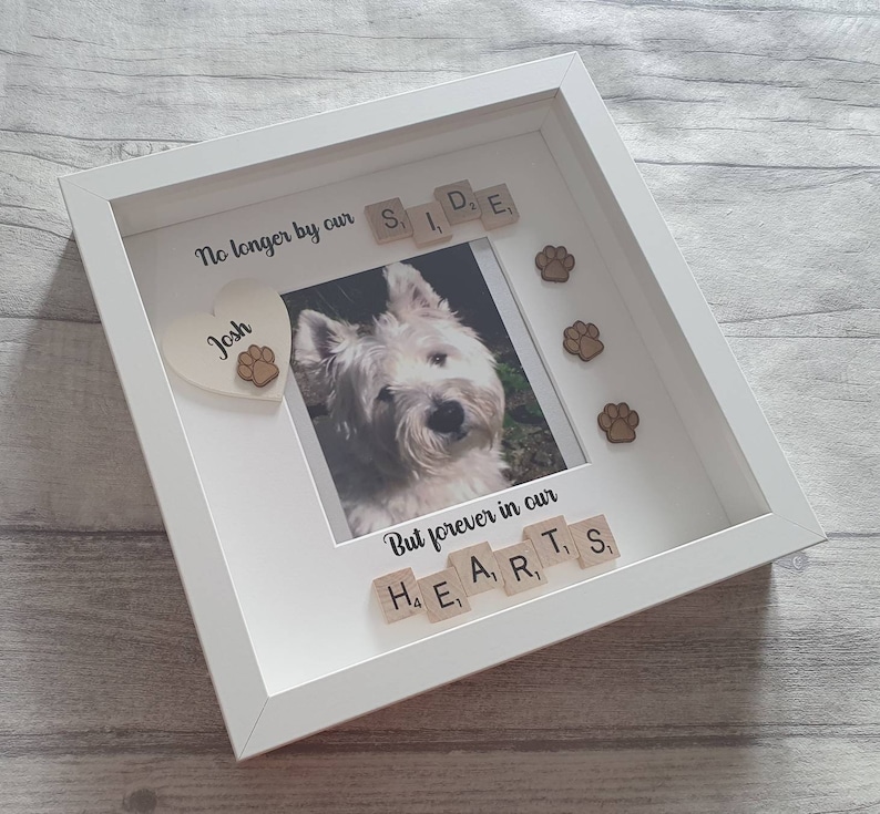 May include: White framed photo memorial with a picture of a white dog. The frame has a heart with the name "Josh" and paw prints. The text "No longer by our side" and "But forever in our hearts" is spelled out with letter tiles.