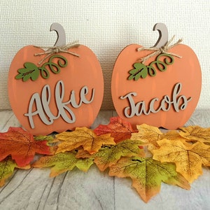 May include: Two orange wooden pumpkin decorations, each with a name in cursive: "Alfie" and "Jacob". They feature beige stems, green leaves, and twine bows. Autumn leaves in shades of red, orange, and yellow are arranged below.