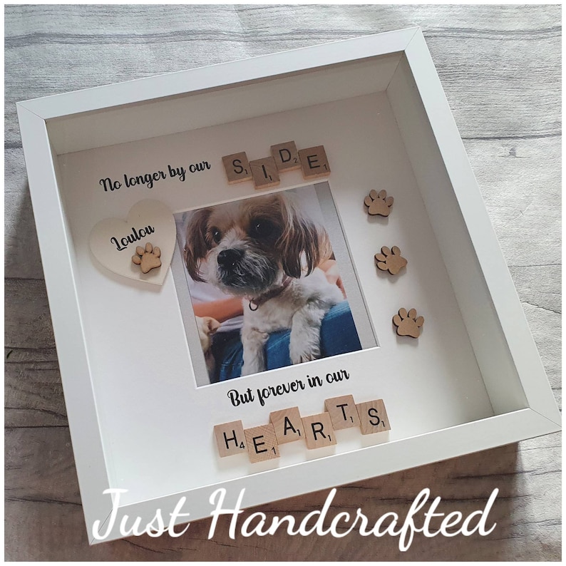 May include: White picture frame with a photo of a dog inside. The frame has a wooden heart with the word "Loulou" and a paw print on it. The frame also has wooden tiles spelling out "No longer by our side" and "But forever in our hearts".