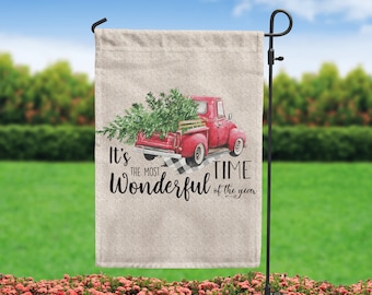 Op de afbeelding: Een rode pick-up truck met een kerstboom op de laadbak, die rijdt op een zwart-wit geruit patroon. De tekst luidt "Het is de mooiste tijd van het jaar".