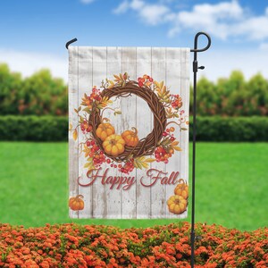 Puede incluir: Una bandera de jard&iacute;n con un dise&ntilde;o r&uacute;stico que presenta una corona de follaje oto&ntilde;al, calabazas y bayas. La bandera tiene un fondo de madera desgastada y las palabras "Happy Fall" en una fuente de escritura. La bandera se muestra en un soporte de metal negro.