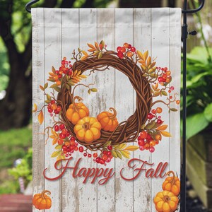 Puede incluir: Una bandera de jard&iacute;n decorativa con tem&aacute;tica oto&ntilde;al. La bandera muestra una corona de ramitas, calabazas naranjas y bayas rojas. Las palabras "Happy Fall" est&aacute;n escritas en rojo sobre un fondo de madera blanca.