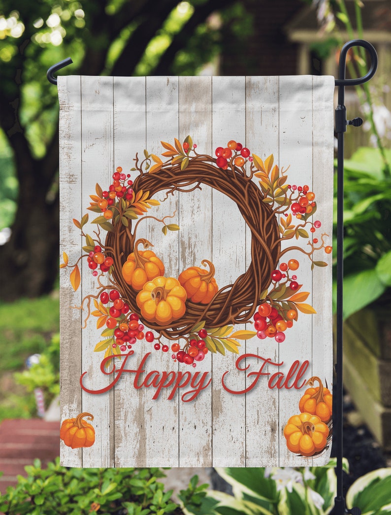 Puede incluir: Una bandera de jard&iacute;n decorativa con tem&aacute;tica oto&ntilde;al. La bandera muestra una corona de ramitas, calabazas naranjas y bayas rojas. Las palabras "Happy Fall" est&aacute;n escritas en rojo sobre un fondo de madera blanca.