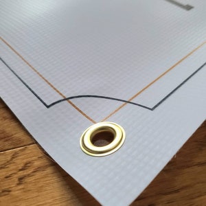 Op de afbeelding: Close-up van een witte banner met een gouden en zwarte rand. De banner heeft een messing oogje in de rechterbenedenhoek.