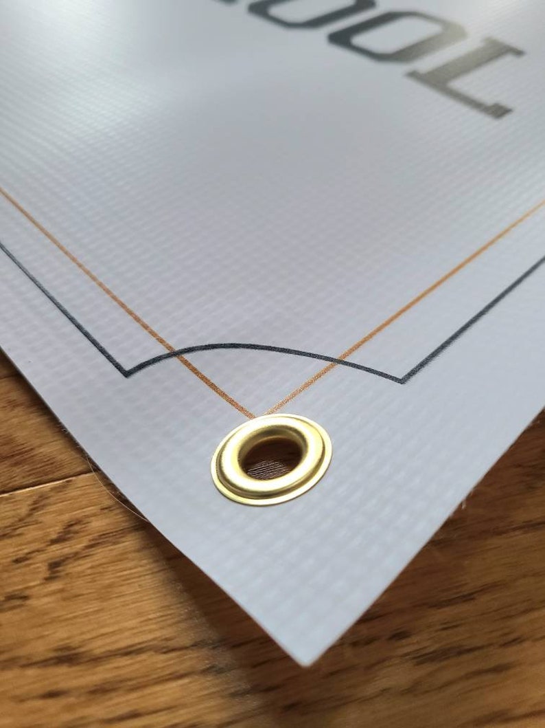Op de afbeelding: Close-up van een witte banner met een gouden en zwarte rand. De banner heeft een messing oogje in de rechterbenedenhoek.