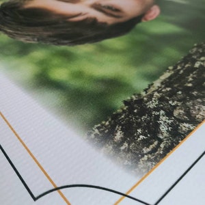 Op de afbeelding: Een close-up van een foto in een wit frame met een gouden en zwarte rand. De foto toont het gezicht van een persoon met gesloten ogen, die omhoog naar de camera kijkt. De achtergrond is wazig en groen.