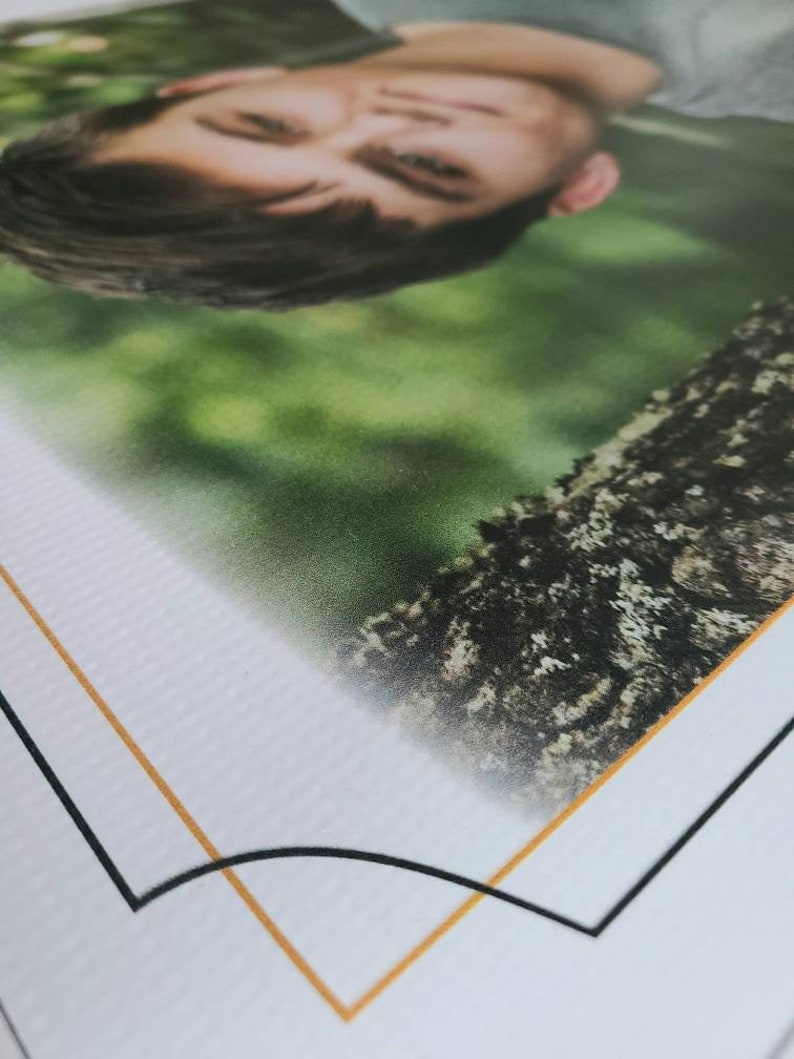 Op de afbeelding: Een close-up van een foto in een wit frame met een gouden en zwarte rand. De foto toont het gezicht van een persoon met gesloten ogen, die omhoog naar de camera kijkt. De achtergrond is wazig en groen.