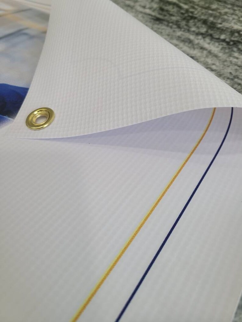 Op de afbeelding: Close-up van een witte banner met een gouden en blauwe naad. De banner heeft een metalen oogje in de linkerbovenhoek.