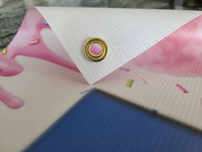 May include: Close-up of a white banner with a gold grommet. The banner is printed with a pink and white design with sprinkles.