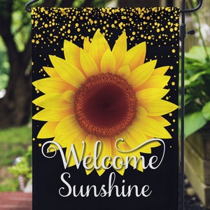 Puede incluir: Una bandera de jard&iacute;n negra con un girasol amarillo y purpurina dorada. El texto "Welcome Sunshine" est&aacute; escrito en letras blancas en cursiva.