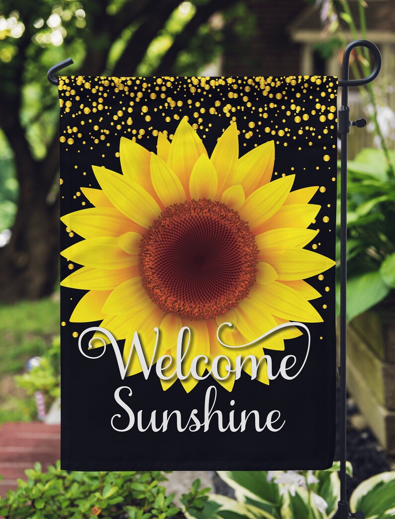 Puede incluir: Una bandera de jard&iacute;n negra con un girasol amarillo y purpurina dorada. El texto "Welcome Sunshine" est&aacute; escrito en letras blancas en cursiva.