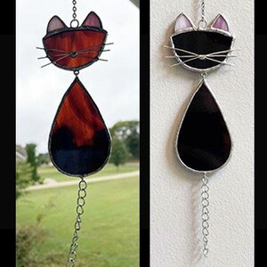 May include: Stained glass cat suncatcher with a black and red color scheme. The cat's head is at the top, with a teardrop-shaped body below. The cat's head has whiskers and ears. A chain is attached to the top for hanging, and a chain hangs from the bottom.