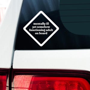 May include: White diamond-shaped sticker with the text "mentally ill yet somehow functioning adult on board" on a black car window.