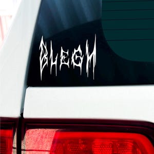以下が含まれることがあります： スタイリッシュなゴシック体で「BLEGH」と書かれた白いビニールステッカー。ステッカーは車のリアウィンドウに貼られています。