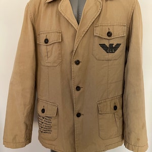 Puede incluir: Una chaqueta beige con botones, cuello, mangas largas y cuatro bolsillos delanteros con cierre de botón. El pecho izquierdo muestra un logotipo de águila negra con las letras GA, mientras que el área del bolsillo inferior derecho presenta texto repetido de Emporio Armani.