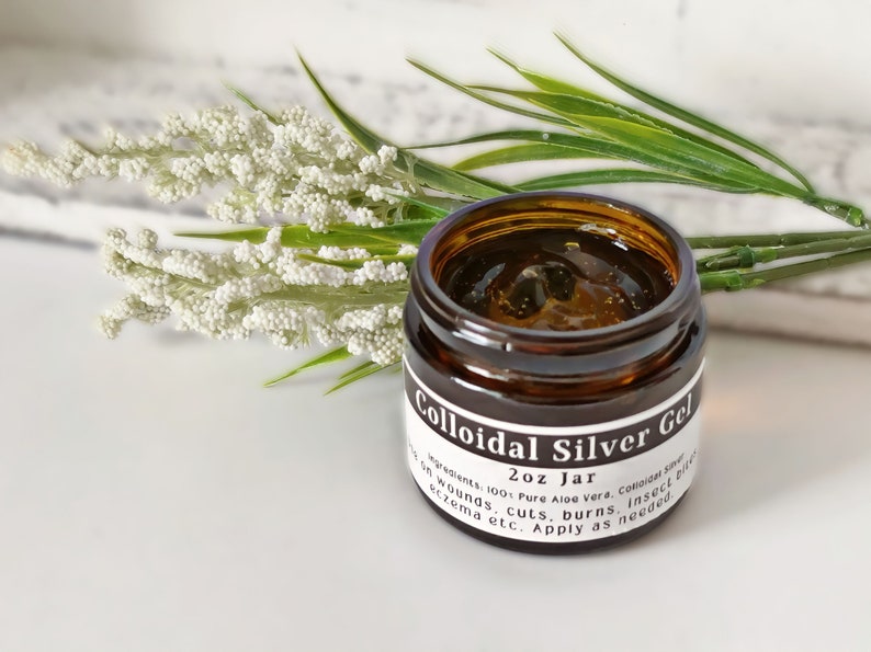 May include: A brown glass jar with a white label that reads "Colloidal Silver Gel 2oz Jar" and lists ingredients and uses for the gel. The jar is sitting on a white surface with a sprig of white flowers and green leaves.
