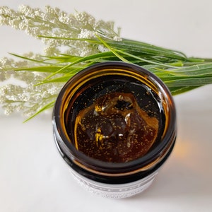 May include: A close-up of a small amber glass jar filled with a clear, golden liquid. The liquid has small, shimmering particles suspended in it. The jar is sitting on a white surface.