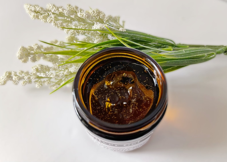 May include: A close-up of a small amber glass jar filled with a clear, golden liquid. The liquid has small, shimmering particles suspended in it. The jar is sitting on a white surface.